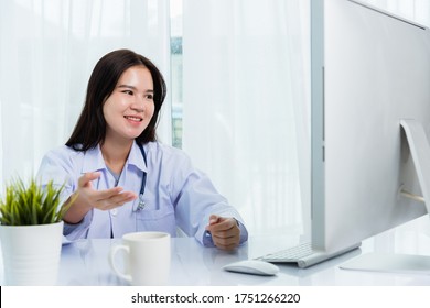 Asian Young Beautiful Woman Doctor Video Call Conference Online Raise Hand To Explain To Patients On Front Desktop PC Computer Monitor Quarantines Disease Coronavirus Or COVID-19 At Hospital Office