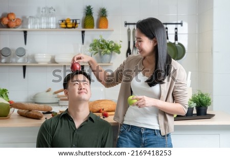 Asian young beautiful woman having fun with place a red apple on head of her handsome man in kitchen while cooking healthy food together. Cute teasing couple in modern kitchen concept