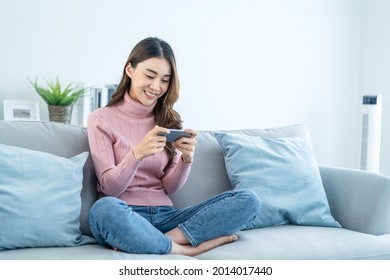 Asian Young Beautiful Woman Playing Mobile Game On Smartphone At Home. Attractive Casual Girl Feel Happy And Relax, Sit On Sofa Having Fun Touching Screen On Phone To Play Video Enjoy Victory In House
