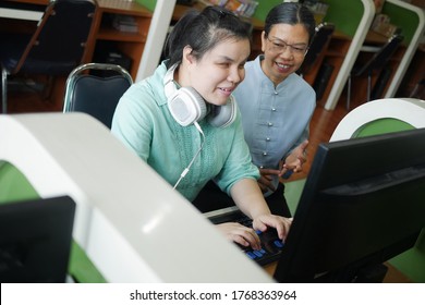 Asian young blind person woman with headphone using computer with braille display assistive device discussing with senior colleague woman in workplace.