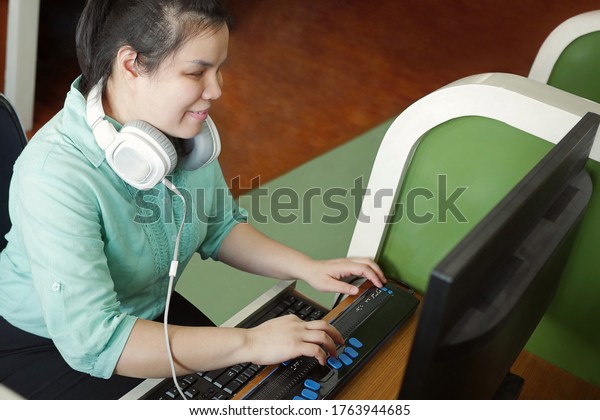 Asian young blind\
person woman with headphone using computer with refreshable braille\
display or braille terminal a technology device for persons with\
visual disabilities.