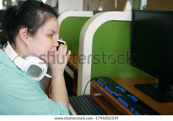 Asian young\
blind person woman with headphone using smart phone with voice\
assistive technology for disabilities persons in workplace with\
computer and braille display on\
table.