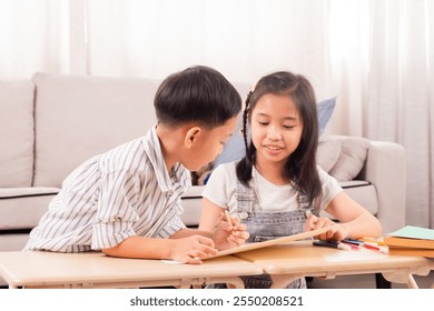 Asian young boy and girl studying together at home. Boy assisting girl with homework, pointing at paper. Cozy living room setting with sofa and stuffed toy in background. Education teamwork. - Powered by Shutterstock