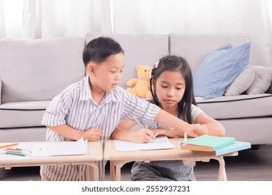 Asian young boy and girl studying together at home. Boy assisting girl with homework, pointing at paper. Cozy living room setting with sofa and stuffed toy in background. Education teamwork. - Powered by Shutterstock