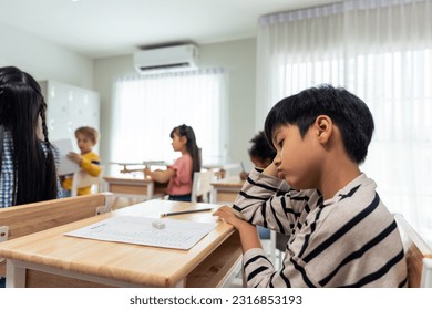 Asian young boy student doing an exam test at elementary school. Adorable children sitting indoors on table, feeling upset and depressed while writing notes, learning with teacher at kindergarten. - Powered by Shutterstock