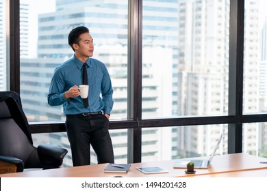 Asian Young Business Man Drinking Coffee Looking Through Window On The Cityscape View . Manager Relaxing In The Office. Thinking . Resting . Take A Break . Alone