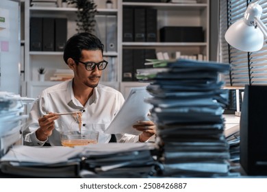 Asian young businessman eating noodles while working in office at night. Attractive professional male employee sitting on table, feeling hungry and starving then having dinner in corporate workplace. - Powered by Shutterstock