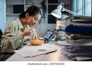 Asian young businesswoman eating noodles while work in office at night. Attractive professional female employee sitting on table, feeling hungry and starving then having dinner in corporate workplace. - Powered by Shutterstock