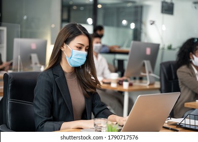 Asian Young Businesswoman Working On Computer In Office With New Normal Lifestyle Concept. Man And Woman Wear Protective Face Mask And Keep Distancing To Prevent Covid Virus After Company Reopen Again