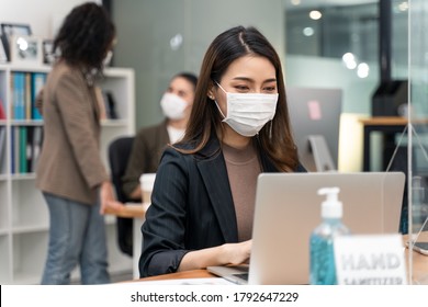 Asian Young Businesswoman Working On Computer In Office With New Normal Lifestyle Concept. Man And Woman Wear Protective Face Mask And Keep Distancing To Prevent Covid Virus After Company Reopen Again