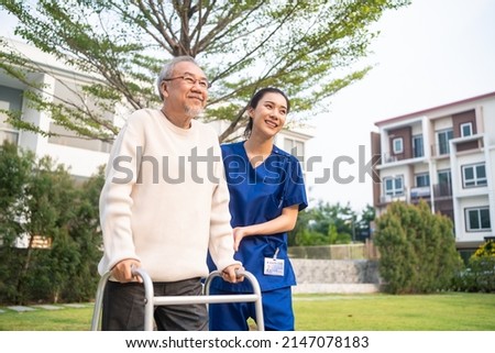 Asian young caregiver nurse support senior older male walking outdoors. Specialist girl doctor help and take care of elderly mature man patient doing physical therapy in public park at nursing home.