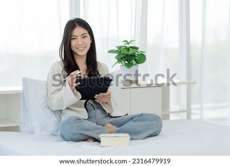 Asian young cheerful female teenage model sitting smiling on white clean sheet bed using pen with touchscreen tablet computer browsing surfing internet shopping working online in modern decor bedroom.