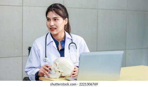 Asian Young Cheerful Happy Professional Female Practitioner Doctor In White Lab Coat Uniform With Stethoscope Sit Smiling Holding Human Skull Skeleton Anatomy Model In Hands At Office Working Desk.