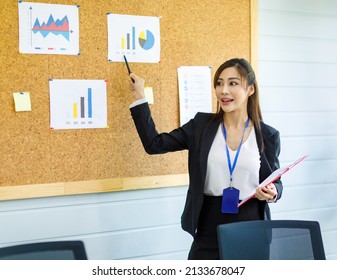 Asian Young Confident Professional Female Businesswoman Employee Staff In Formal Suit Holding Clipboard And Pen Pointing Presenting Sales Target Graph Chart Diagram Paperwork On Board In Meeting Room.