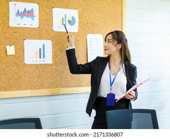 Asian Young Confident Professional Female Businesswoman Employee Staff In Formal Suit Holding Clipboard And Pen Pointing Presenting Sales Target Graph Chart Diagram Paperwork On Board In Meeting Room.