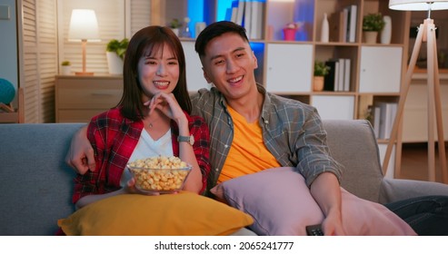 Asian Young Couple Eating Pop Corn Are Watching Tv And Sitting On The Couch Happily In Night Living Room At Home