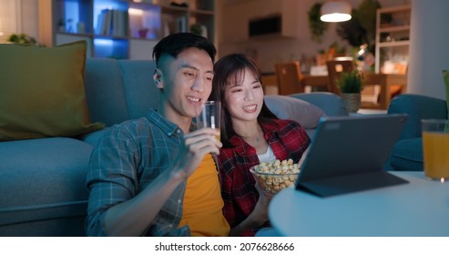asian young couple eating pop corn are watching movie or video by digital tablet and sitting on the couch happily in night living room at home
