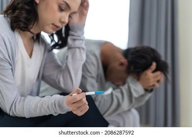 Asian Young Couple Feeling Shock After Look At Positive Pregnancy Test. Disappointed Unplanned Pregnancy Men And Women Sit On Bed In Bedroom With Upset And Sad Face For Problem Don't Want To Pregnant.