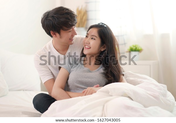 Asian young couple man and woman staying\
together in bedroom. Husband hugging wife from behind, looking eyes\
at each other feeling lover happy with smile face on morning.\
Relationship of love\
concept.