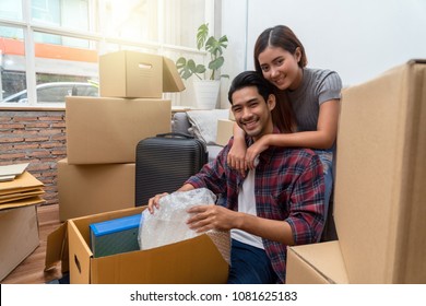 Asian Young Couple Packing Into Big Cardboard Box For Moving In New Home, Sitting On Difference Step Between Sofa And Floor And Hug At The Relax Time, Moving And House Hunting Concept