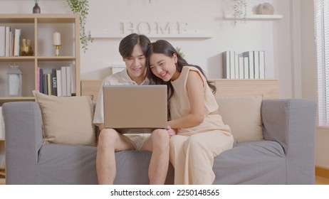 Asian young couple sit on couch or sofa looking at Computer Laptop smile and laughing together. Cheerful lover surfing internet and social media spending time in holiday at cozy home. Valentine Day - Powered by Shutterstock