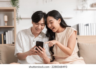 Asian young couple sit on couch or sofa looking at mobile phone smile and laughing together. Cheerful lover surfing internet and social media spending time in holiday at cozy home. Valentine Day