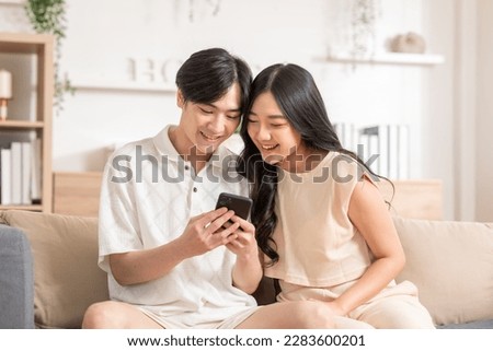 Asian young couple sit on couch or sofa looking at mobile phone smile and laughing together. Cheerful lover surfing internet and social media spending time in holiday at cozy home. Valentine Day