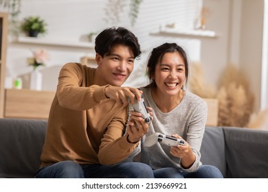 Asian young couple sitting on couch in living room enjoy playing video games spending time together. Happy couple play video games at home both are smiling, laughing and enjoying moment on weekend.