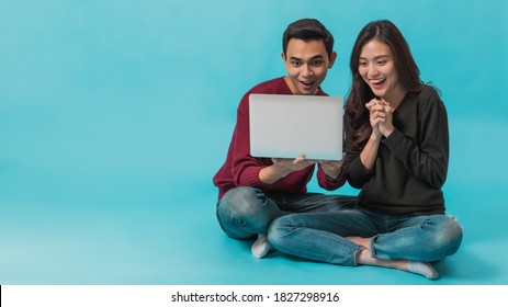 Asian young couple sitting and using laptop with smiling face together isolated on blue background.Concept of happy couple working with online technology.