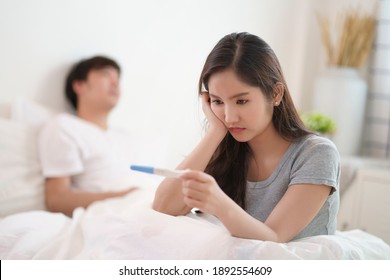 Asian young couple wife and husband find out a results of a pregnancy test in bedroom at home. Sad woman looking at pregnancy test. Husband upset and sit on the bed and unhappy feeling.