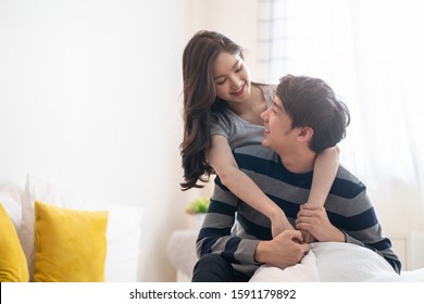 Asian Young Couples Man And Woman Staying Together In Bed Room. Girl Hugging Boy From Behind, Looking Eyes At Each Other Feeling Love Happy With Smile Face. Relationship Of Young Girl And Boy Concept