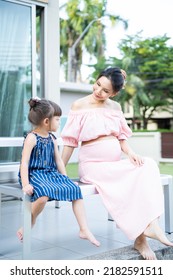 Asian Young Daughter Talk With Loving Beautiful Pregnant Mom Outdoors. Attractive Expecting Female Pregnancy Embrace Little Kid Girl With Happiness And Love While Sitting Outside In Backyard At Home.