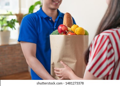 Asian Young Delivery Man In Blue Shirt Uniform Delivering Package Bag Fresh Food To Woman At Home.Grocery Store Delivery Service.