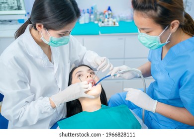 Asian Young Dentist Wearing Face Mask Use Medical Instruments For Oral Care Treatment. Assistant Sitting Beside Using Suction Machine To Keep Patient's Mouth Dry During Treatment Procedures At Clinic.