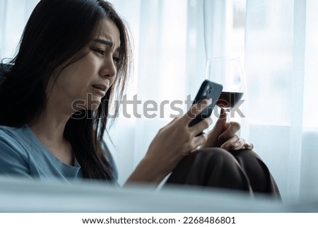 Asian young depression woman drinking wine while look at mobile phone. Attractive beautiful girl sitting on floor hold a bottle of alcohol feeling heart broken, drunk and hangover alone in living room