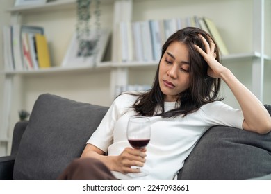 Asian young depression woman feeling heart broken and drinking alcohol. Attractive beautiful girl feel loneliness, drunk and hangover alone, lying down on sofa, holding a glass of wine in living room.