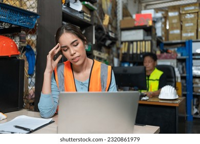 Asian young engineer female feeling stress and headache while working with laptop at warehouse factory. Woman worker in vest safety working in store office. - Shutterstock ID 2320861979