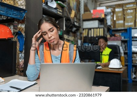 Asian young engineer female feeling stress and headache while working with laptop at warehouse factory. Woman worker in vest safety working in store office.