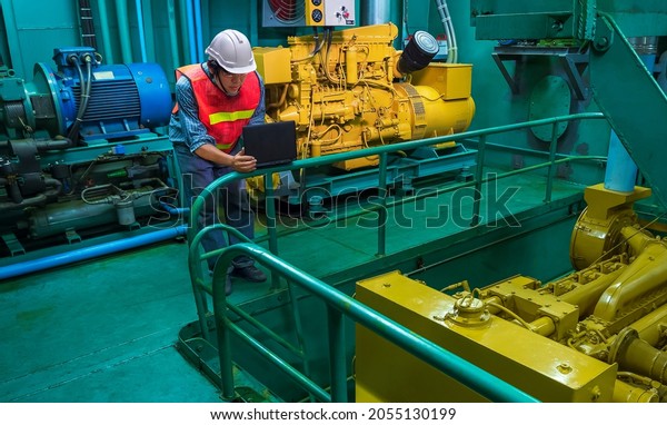 Asian young engineer in reflective\
clothing with helmet using laptop computer to checking engine\
quality system inside of engine room of fishing\
vessel