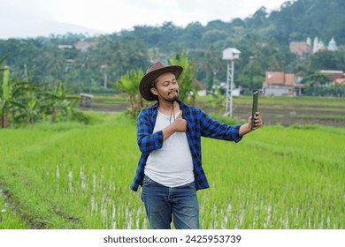 Asian young farmer using tablet at the green rice field. Happy Asian farmers video call with family by smartphone at organic rice field. Video call with tablet. 