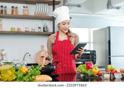 Asian young female chef housewife wears white tall cook hat and apron learning cooking homemade food meal online via touchscreen tablet computer while using spatula frying eggs in pan at home kitchen. - Powered by Shutterstock