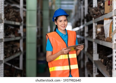 Asian young female employee warehouse worker in safety vest and blue helmet using tablet for checking products or barcode symbol on parcel spare parts on shelf pallet in industry factory warehouse