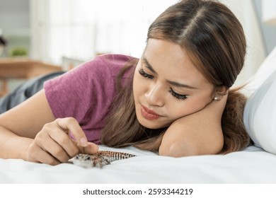 Dueña joven asiática juega con su gecko leopardo en el dormitorio en casa. Mujer atractiva acostada en la cama, pasar tiempo de ocio con el lagarto doméstico en la casa. Mascotas amigables y concepto de interacción