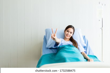 Asian Young Female Patient Smiley Face Lifts Two Fingers Up Fighting With Illness On Bed In The Room Hospital Background.