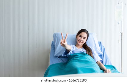 Asian Young Female Patient Smiley Face Lifts Two Fingers Up Fighting With Illness On Bed In The Room Hospital Background.