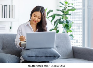 Asian young female professional successful businesswoman in casual outfit sitting on cozy sofa using pen writing note in notebook while working with laptop computer at home office in living room. - Powered by Shutterstock