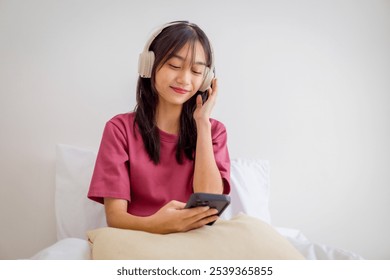 Asian young girl listening music with wireless headphones while sitting on bed. Teenage woman using mobile phone turn on music playlist and enjoy morning activities relaxing in bedroom at home. - Powered by Shutterstock