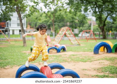 Asian young girl is playing in a park with a tire obstacle course. The park is filled with various play equipment, including swings and a slide. The atmosphere is lively and fun - Powered by Shutterstock