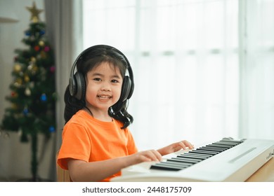 Asian young girl is smiling sitting at a piano, wearing headphones. She is wearing an orange shirt. The room is filled with books and a keyboard - Powered by Shutterstock