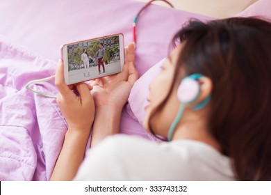 Asian young girl watching mobile video smart phone wear headphones lying on bed